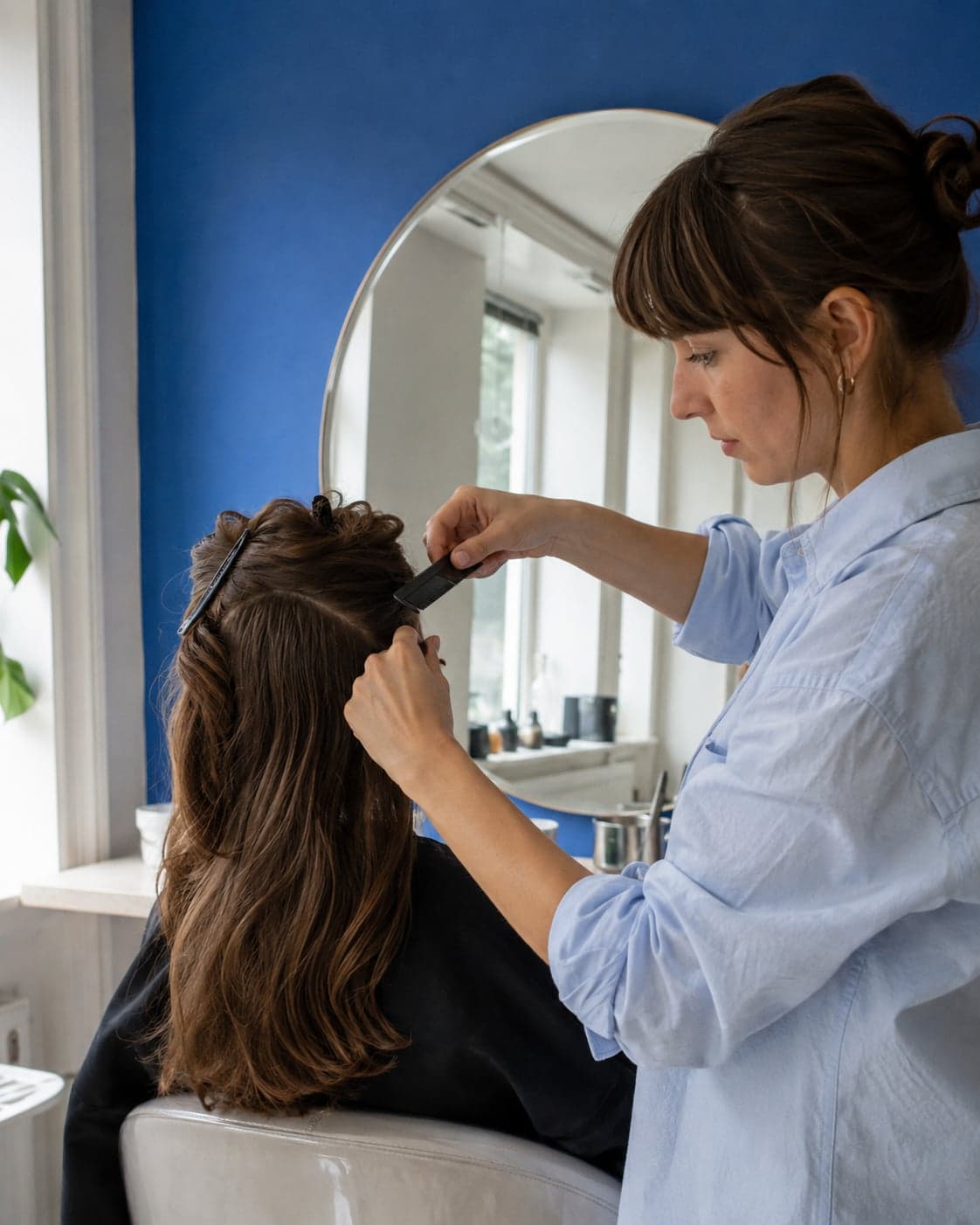 A stylist carefully sections a client’s hair beside a blue salon wall and an arched mirror